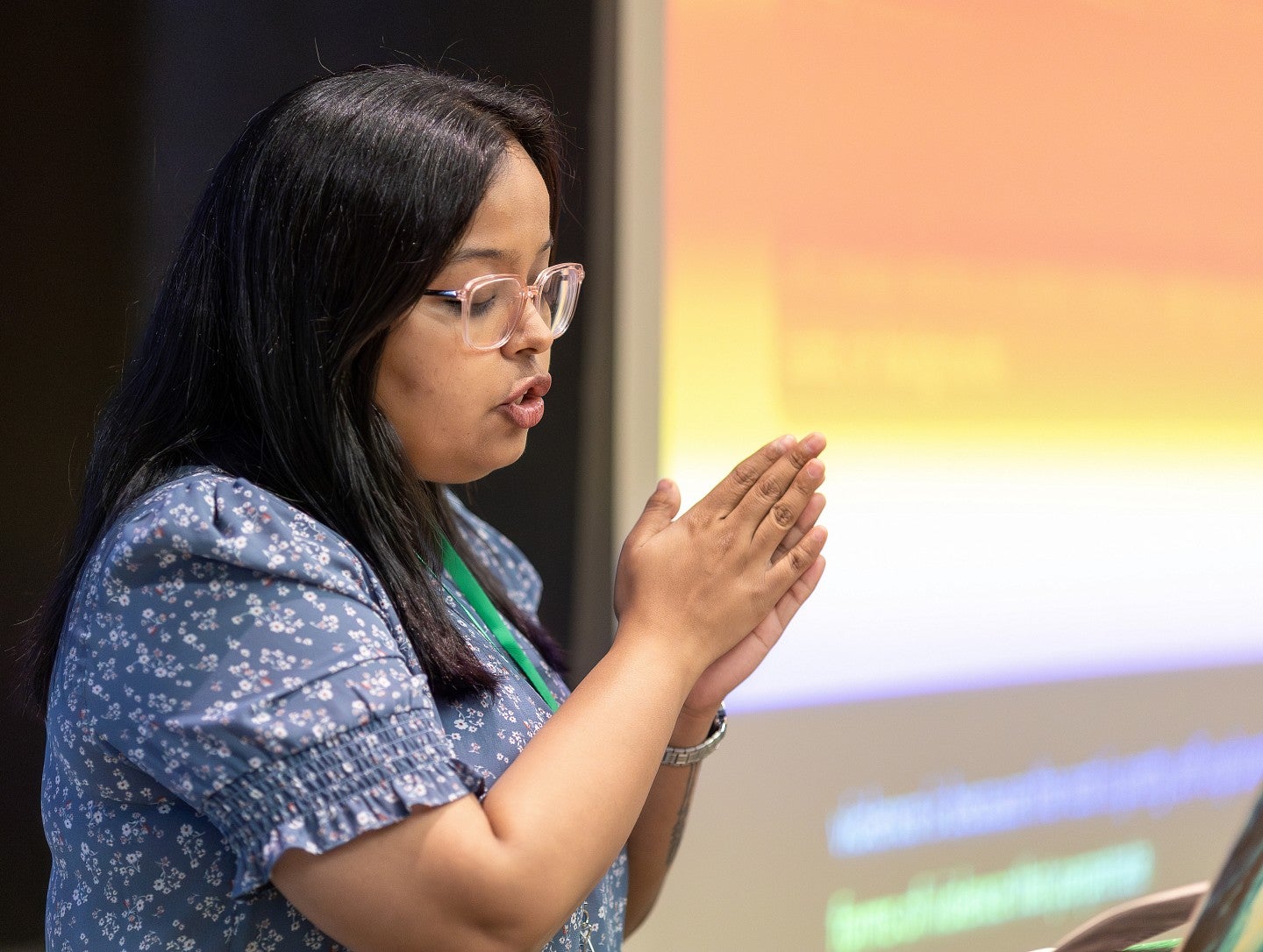 A presenter in front of their slide, mid presentation, hands clasped together.