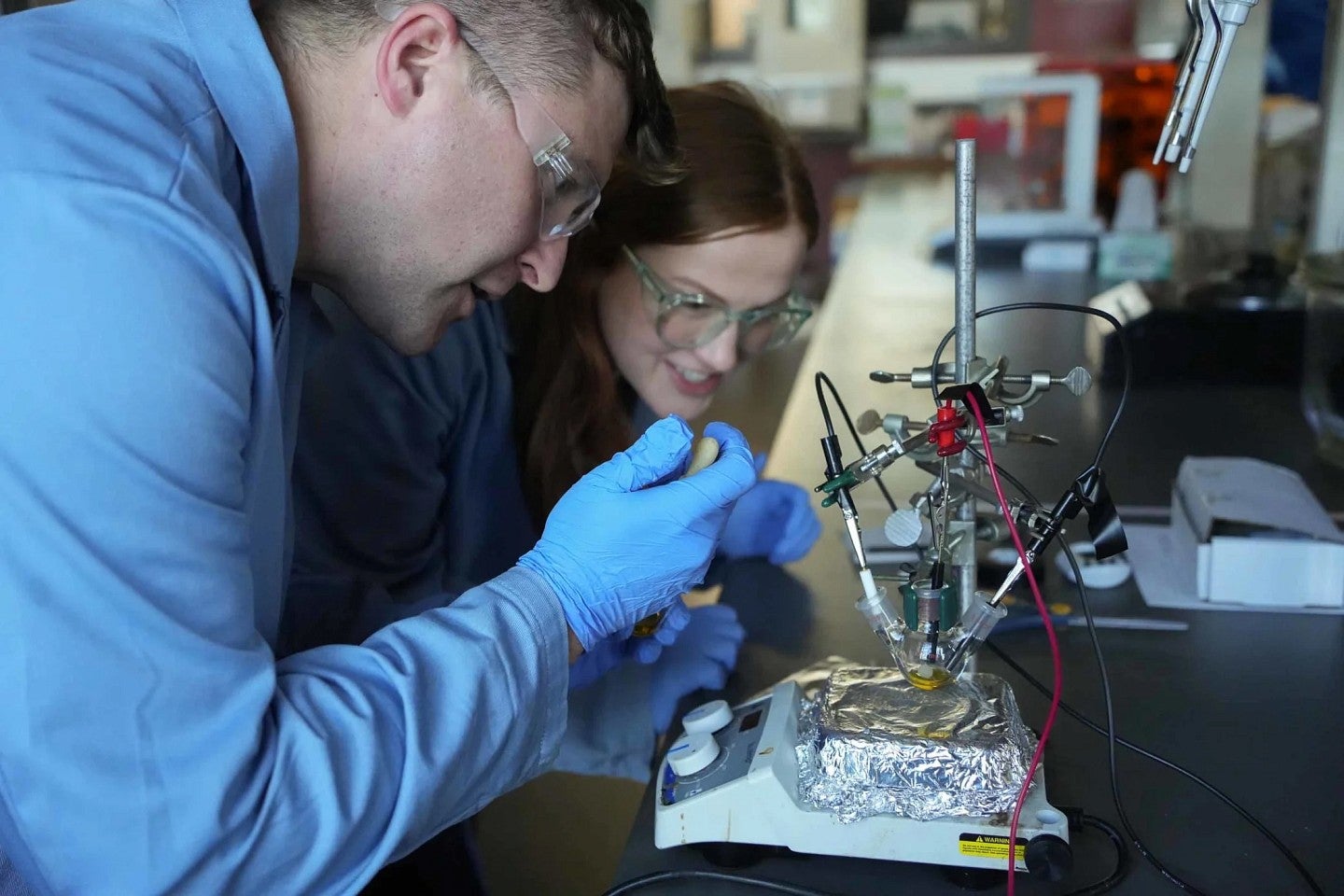 Two scientists wearing safety goggles lean down while conducting an experiment.