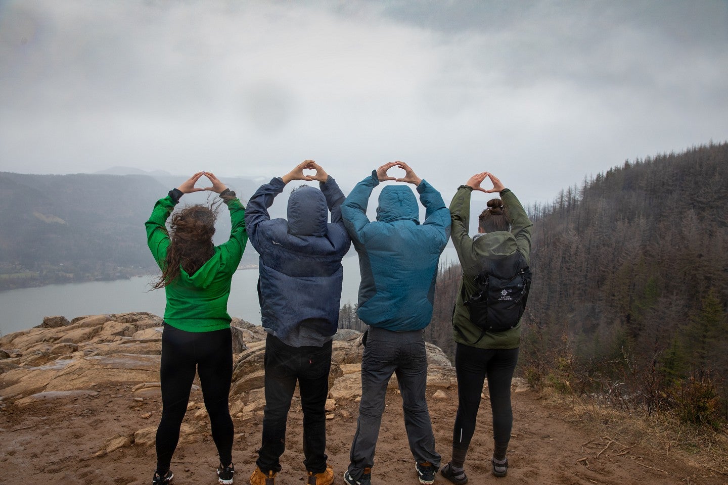 Four students wearing jackets, as seen from behind. They have their hands over their heads and are making the letter "O" with their fingers.