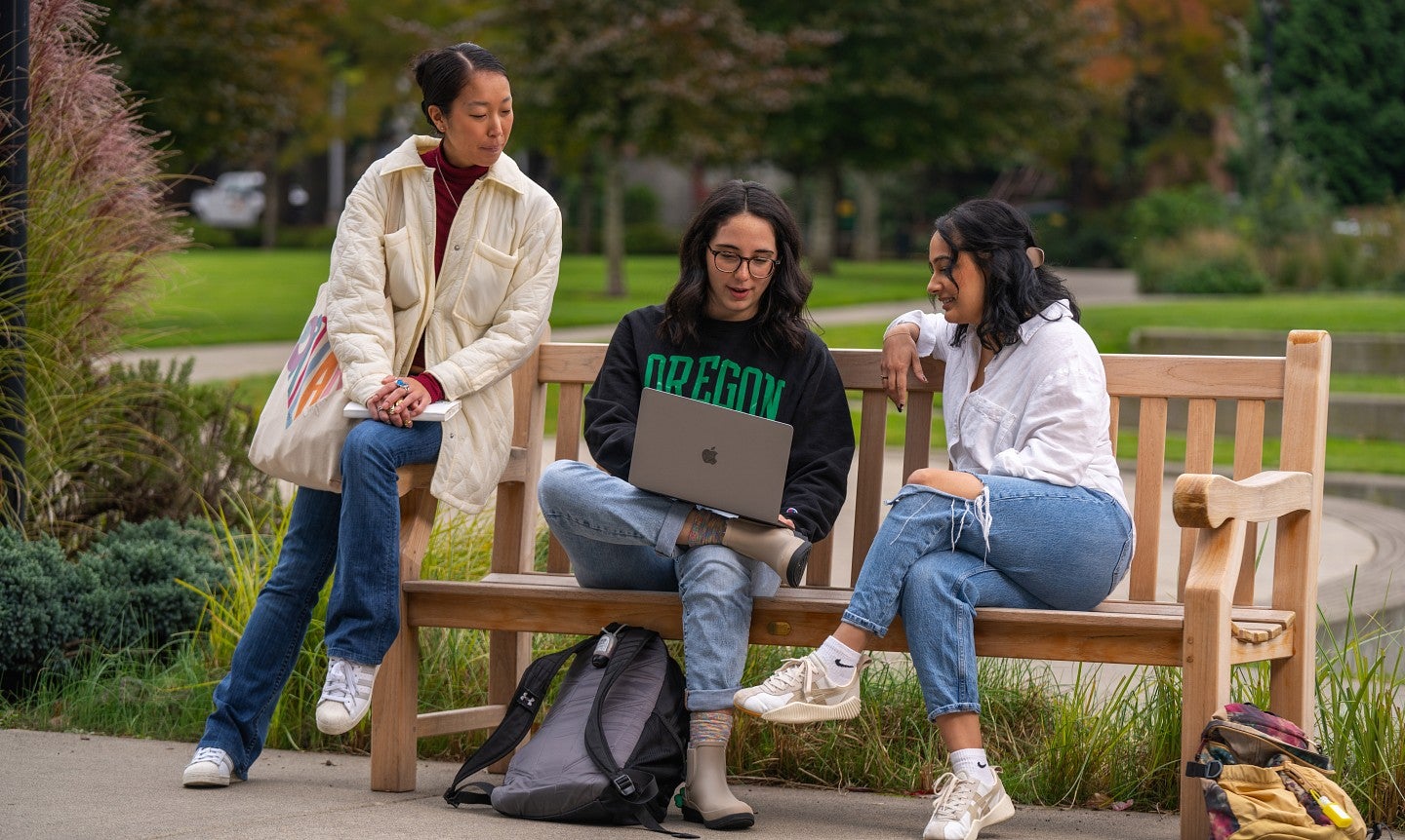 Three graduate students sit on a bench and look together at a laptop.
