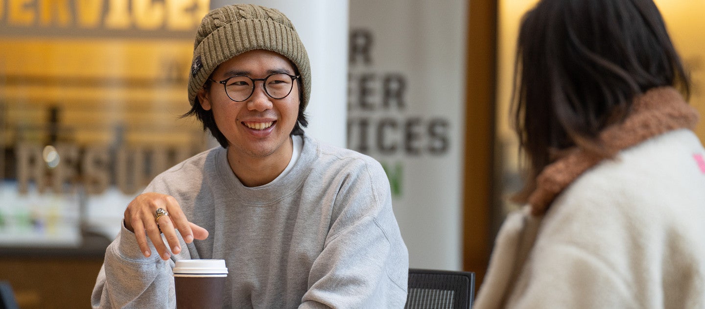 A student in a beanie smiles while drinking coffee.
