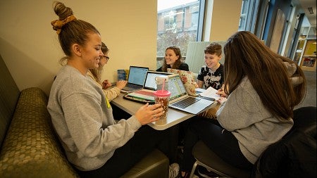 A group of students gather at a small table, using their laptops and having conversation.