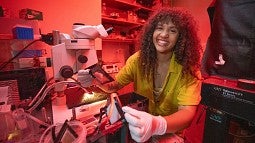 In a lab bathed in red light, Angelique smiles at the camera while using a microscope.