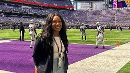 Naomi, a black woman with long, dark hair, smiles while standing on the Minnesota Vikings' field.