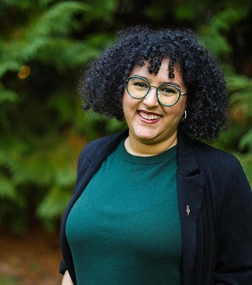 Nahla Bendefaa has dark curly hair and is wearing green framed glasses and red lipstick.