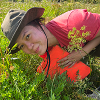 Jasmin, wearing a red shirt and brown hat, leans into tall grass and looks at the camera.