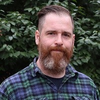 A white man with a beard and cropped hair wearing a checked shirt in front of foliage