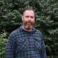A white man with a beard and cropped hair wearing a checked shirt in front of foliage